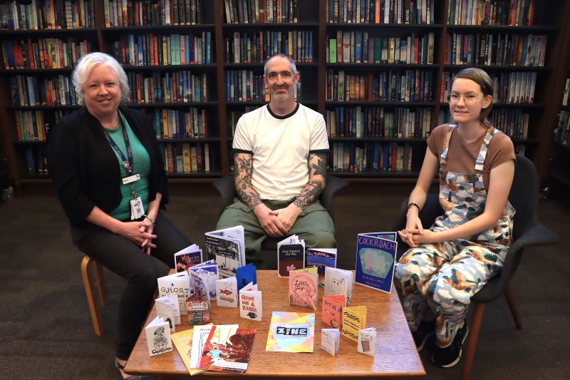 City of Newcastle Innovative Collection Developer Emma Callaghan sits alongside James Turvey and Laura Baker with their zines to be featured in the new collection at Newcastle City Library.