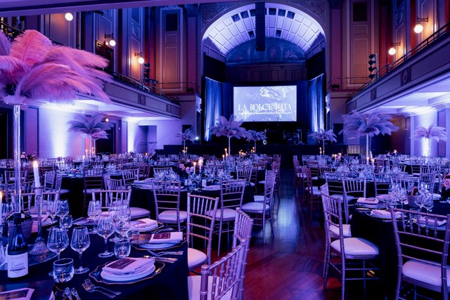 The Concert Hall at Newcastle City Hall set up for a social banquet dinner event, with round tables, feathered decorative table arrangements and purple mood lighting throughout the room.